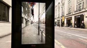 An augmented reality (AR) bus stop advertisement showing a realistic overlay of a person being lifted into the air by balloons, blending seamlessly with the real-world street view in the background.