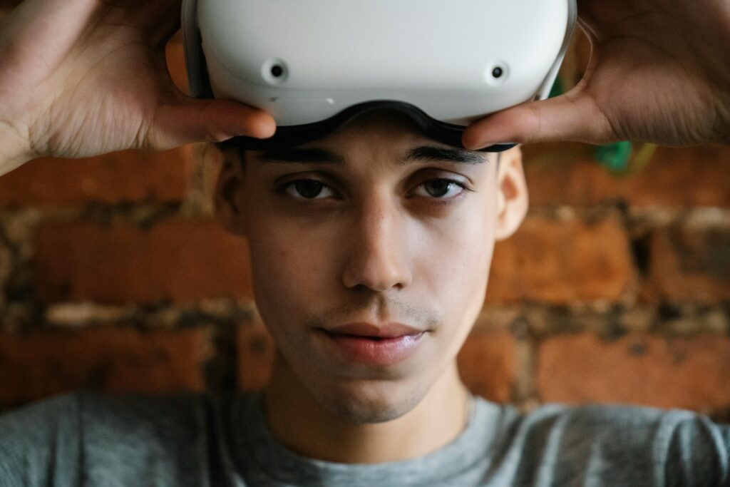 A close-up of a person holding a white virtual reality (VR) headset on their forehead, with both hands adjusting the device. The individual is looking directly at the camera, set against a blurred brick wall background.